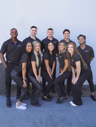 Ten-person smiling staff team in matching black polo shirts and black pants posing outdoors against a light gray wall — upbeat office team portrait.