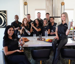 Cheerful cocktail team in matching black polos posing behind a modern marble kitchen island and home bar, holding bottles, shakers, glassware and a platter of croissants and fruit.