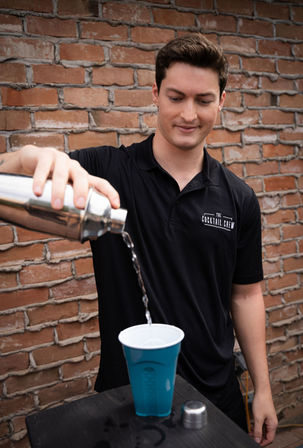 Bartender pouring a cocktail from a stainless steel shaker into a blue plastic cup against a weathered brick wall