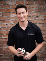 Smiling young bartender holding a stainless steel cocktail shaker in front of a red brick wall for a casual outdoor portrait.
