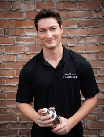Smiling young bartender holding a stainless steel cocktail shaker in front of a red brick wall for a casual outdoor portrait.