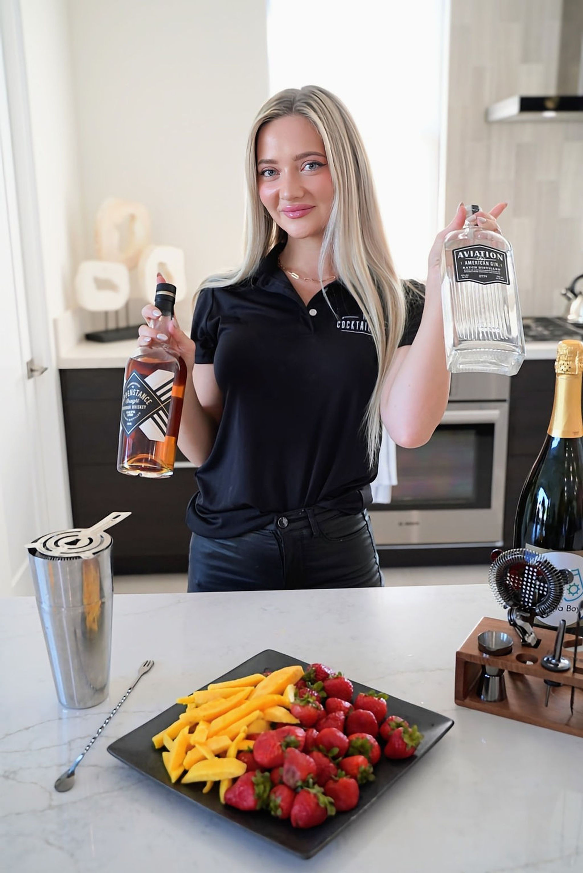 Smiling bartender in a modern kitchen holding a gin bottle and a whiskey bottle over a marble island with cocktail shaker, bar tools, and a plate of strawberries and mango slices — home bar cocktail prep.