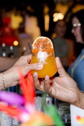 Two hands exchanging a bright orange cocktail in a ribbed glass topped with a dried citrus wheel at a lively bar