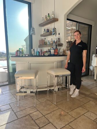 Woman standing beside a modern home bar with two white cushioned stools, bottles on wooden floating shelves, mosaic pendant lights, sunlight streaming through sliding glass doors to a pool and distant hills, tiled floor.