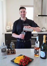 Smiling bartender pouring a pink cocktail from a shaker into a glass at a modern kitchen island bar, with gin, whiskey and sparkling wine bottles and a plate of strawberries and mango slices in the foreground.