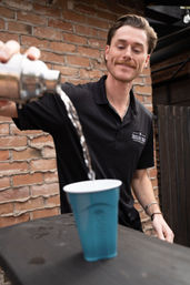 Bartender pouring a clear cocktail from a metal shaker into a blue plastic cup on an urban brick-walled outdoor patio