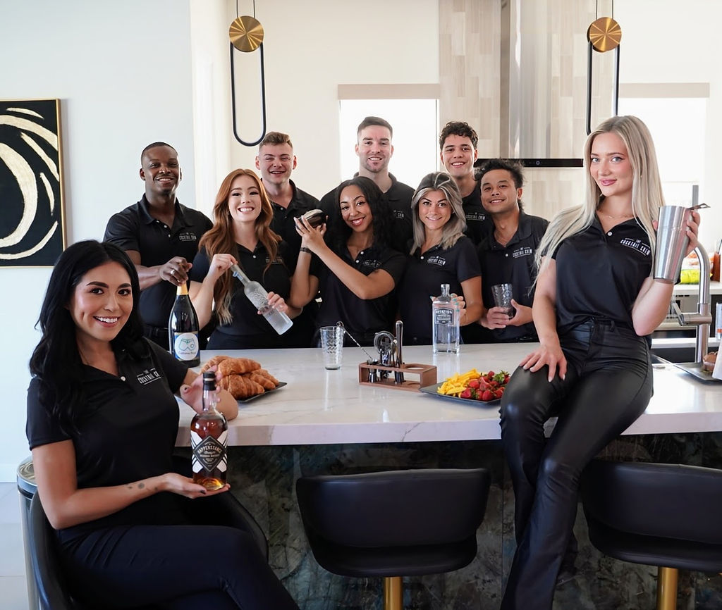 Smiling hospitality team in matching black polos posing at a modern kitchen island with cocktail shakers, liquor bottles, croissants and a fruit platter — casual bar staff group photo