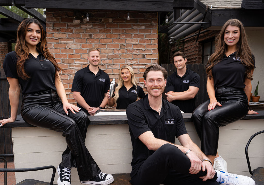 Cheerful crew of six in matching black polos posing at an outdoor patio bar with a brick wall backdrop — two women sitting on the bar and one holding a cocktail shaker, upbeat hospitality team vibe.