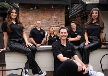 Six-person bar team in black polos posing on a brick-walled outdoor patio — two women seated on the bar counter in leather pants, three men standing behind and one man sitting smiling in front, one teammate holding a cocktail shaker.
