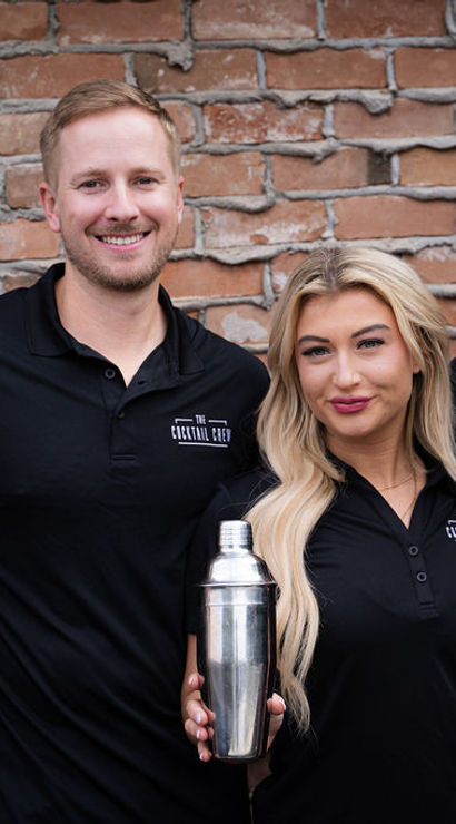 Four bartenders in matching black polos smiling and holding a stainless-steel cocktail shaker in front of an exposed brick wall — urban mixology team portrait.