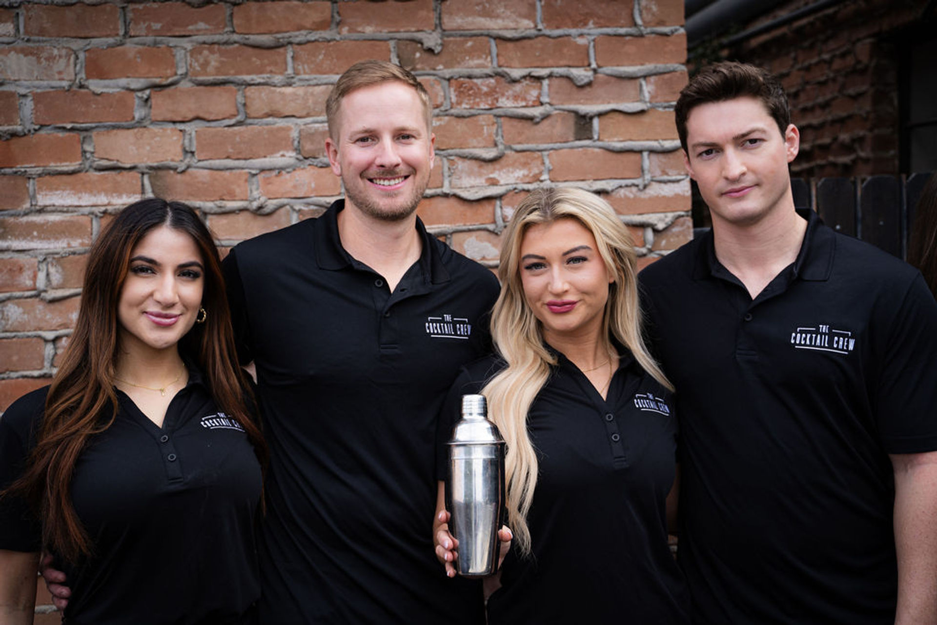 Four bartenders in matching black polos smiling and holding a stainless-steel cocktail shaker in front of an exposed brick wall — urban mixology team portrait.