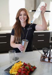 Smiling bartender in a modern kitchen pouring clear liquor into a stainless-steel cocktail shaker, with cocktail tools and a platter of strawberries and mango slices on a marble countertop.