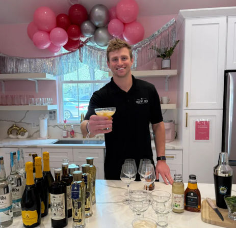 Smiling bartender offers a cocktail in a decorated pink kitchen bar, marble counter lined with champagne and liquor bottles, glassware and a shaker, with a pink and silver balloon garland overhead.