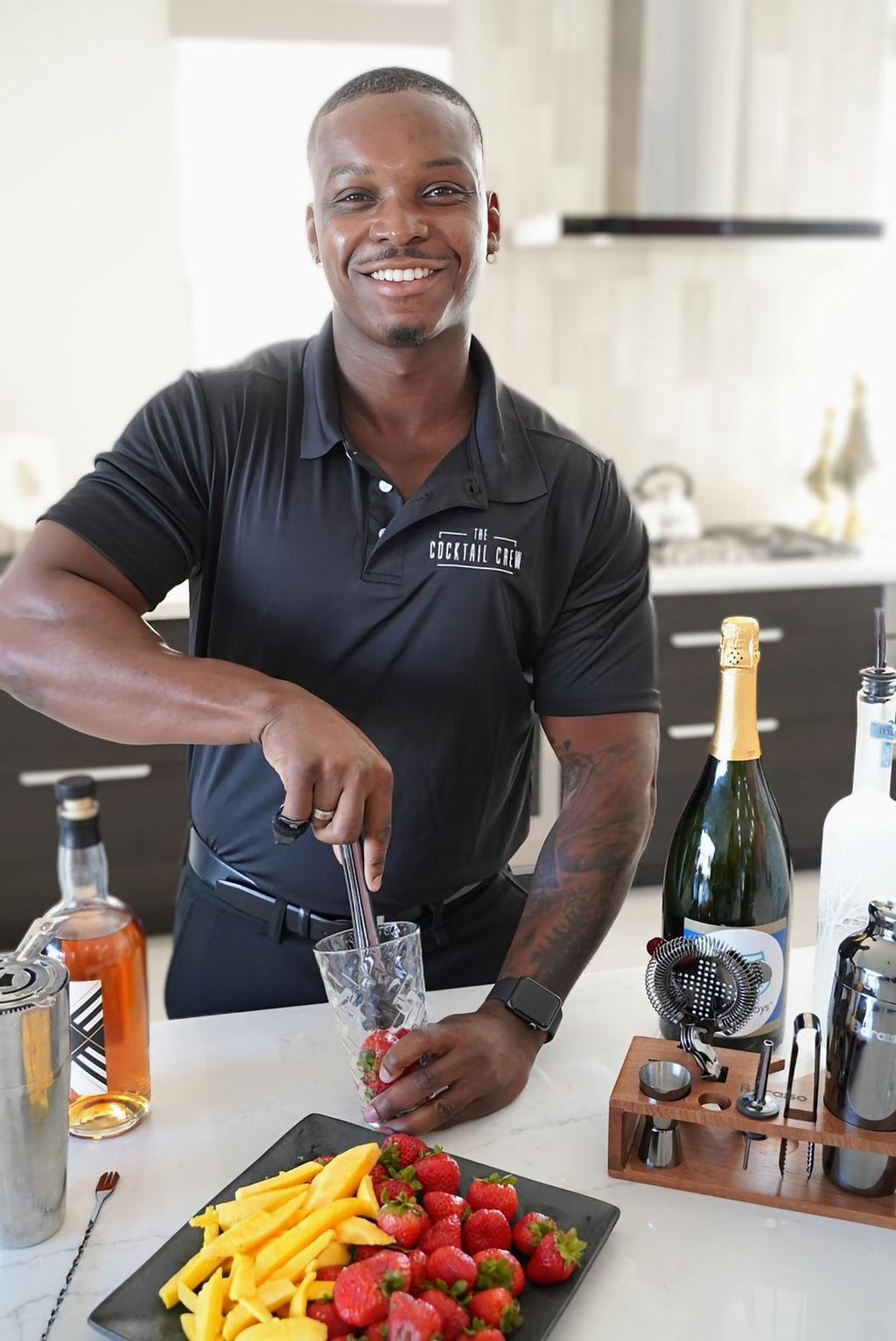 Smiling bartender in a black polo muddling strawberries for a cocktail at a bright modern kitchen island, with mango slices, champagne and bar tools nearby.