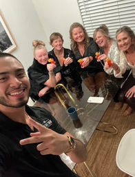 Group selfie of friends — five women and one man — smiling and toasting small red cocktails with orange garnishes around a glass dining table at an indoor gathering.