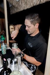 Bartender pouring spirits into a cocktail shaker at a tiki-style outdoor bar at night, preparing mixed drinks with jiggers and glassware.