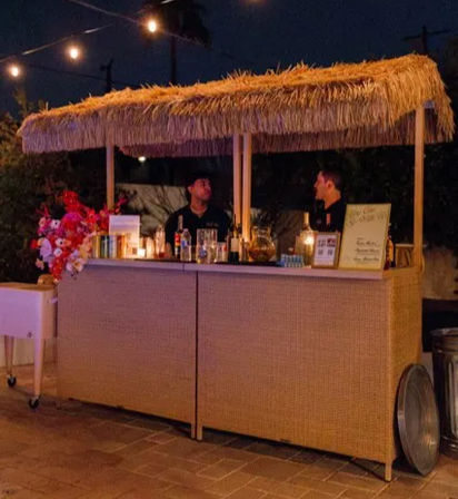 Nighttime outdoor tiki-style bar on a patio with a straw-thatched roof, string lights, woven front counter, two staff, bottles, floral arrangement, menu board and candlelit event vibe