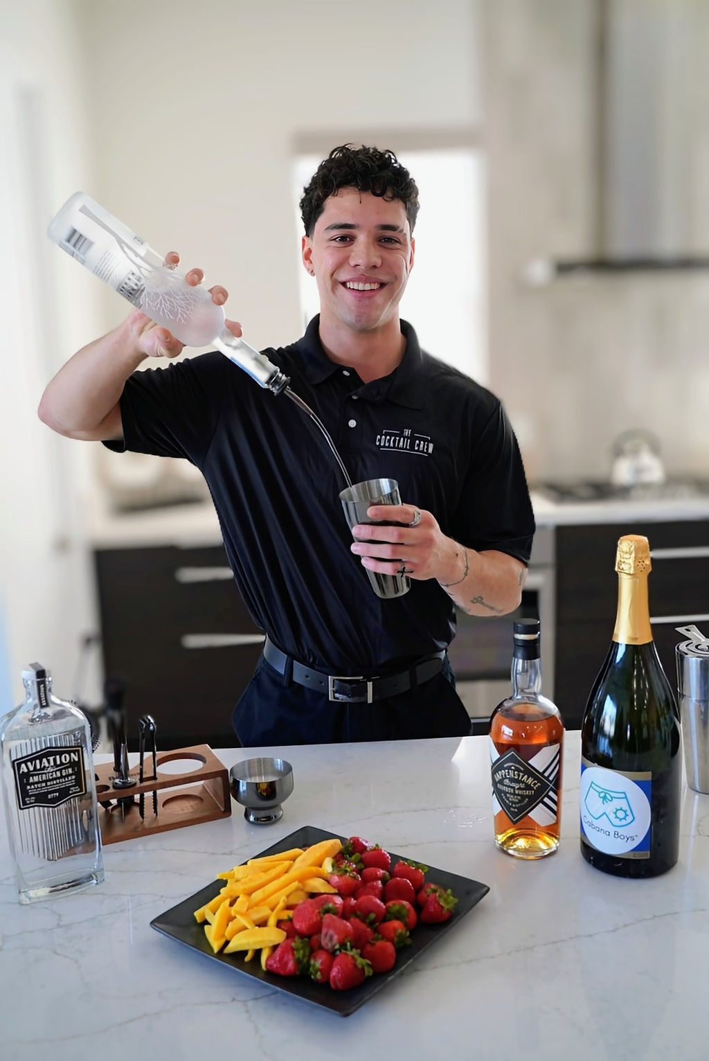 Smiling bartender in a modern kitchen pouring clear liquor into a metal shaker, with whiskey and champagne bottles and a plate of strawberries and mango slices on the counter.