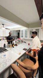 Casual cocktail night in a modern white home kitchen: friends gathered at a marble island while one person prepares drinks amid liquor bottles, glassware and a copper mug.