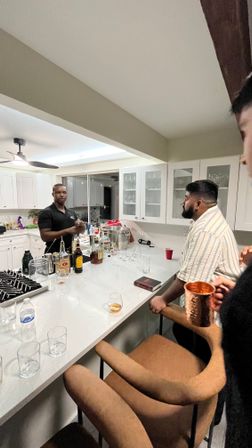 Casual cocktail night in a modern white home kitchen: friends gathered at a marble island while one person prepares drinks amid liquor bottles, glassware and a copper mug.