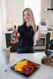 Smiling woman in a modern home kitchen holding two liquor bottles over a counter with a shaker, cocktail tools, and a platter of mango slices and strawberries.