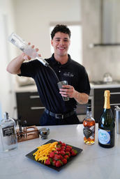 Smiling bartender in a black shirt pouring clear spirit into a metal shaker in a modern kitchen, marble countertop lined with vodka, whiskey and sparkling wine bottles and a plate of strawberries and mango slices.