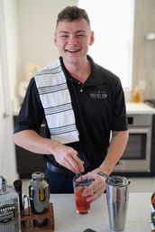Smiling bartender in a black polo with a striped towel over his shoulder stirring a fruit cocktail with a bar spoon at a modern home kitchen bar, cocktail shaker and spirits on the counter.