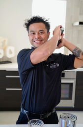 Smiling bartender in a black polo shaking a cocktail over a modern kitchen island, mixing glass and shaker in the foreground