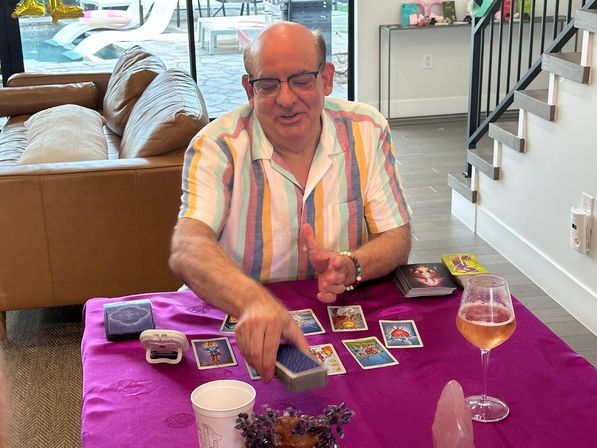 Man performing a tarot card reading at a purple-covered table in a modern living room with crystals, tarot cards, a wine glass, and a pool visible through sliding glass doors.