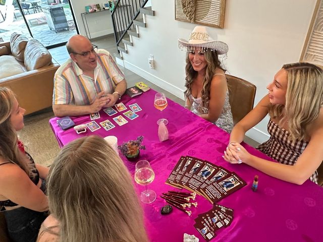 At-home bachelorette tarot reading: friends gathered around a magenta tablecloth with tarot cards, crystals, wine glasses, and a smiling woman in a sequined dress wearing a white beaded BRIDE hat.
