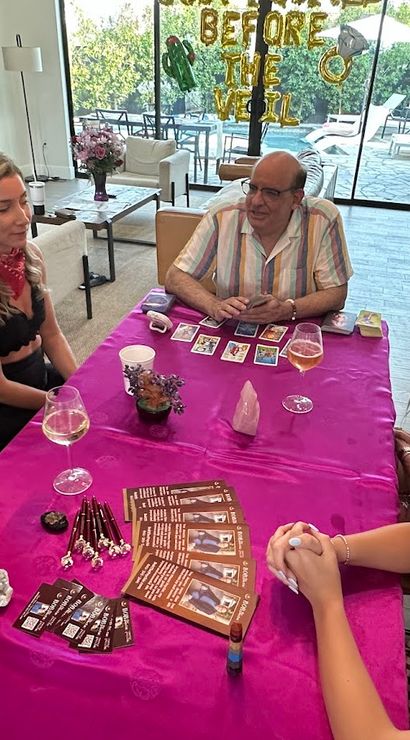 Five people gathered around a magenta-covered table in a bright living room for a tarot card reading and bridal-shower-style celebration, with crystals, tarot cards, wine glasses and a ‘Before the Veil’ balloon banner and patio doors visible
