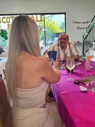 Long-haired woman holding tarot-style cards for a smiling man across a purple-covered table with wine glasses, crystals and party balloons in a bright home living room.