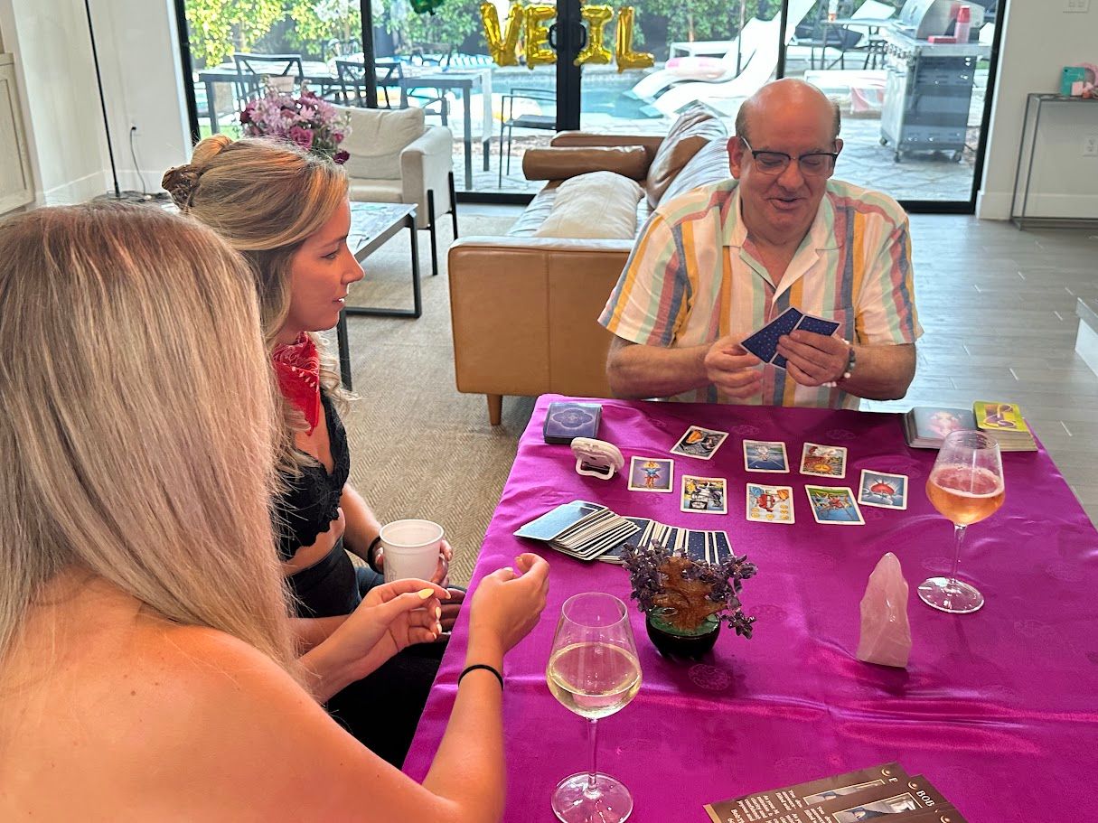 Three adults gathered around a purple-covered table in a bright living room for a tarot card reading, with a tarot spread, reader holding cards, wine glasses and a pink crystal