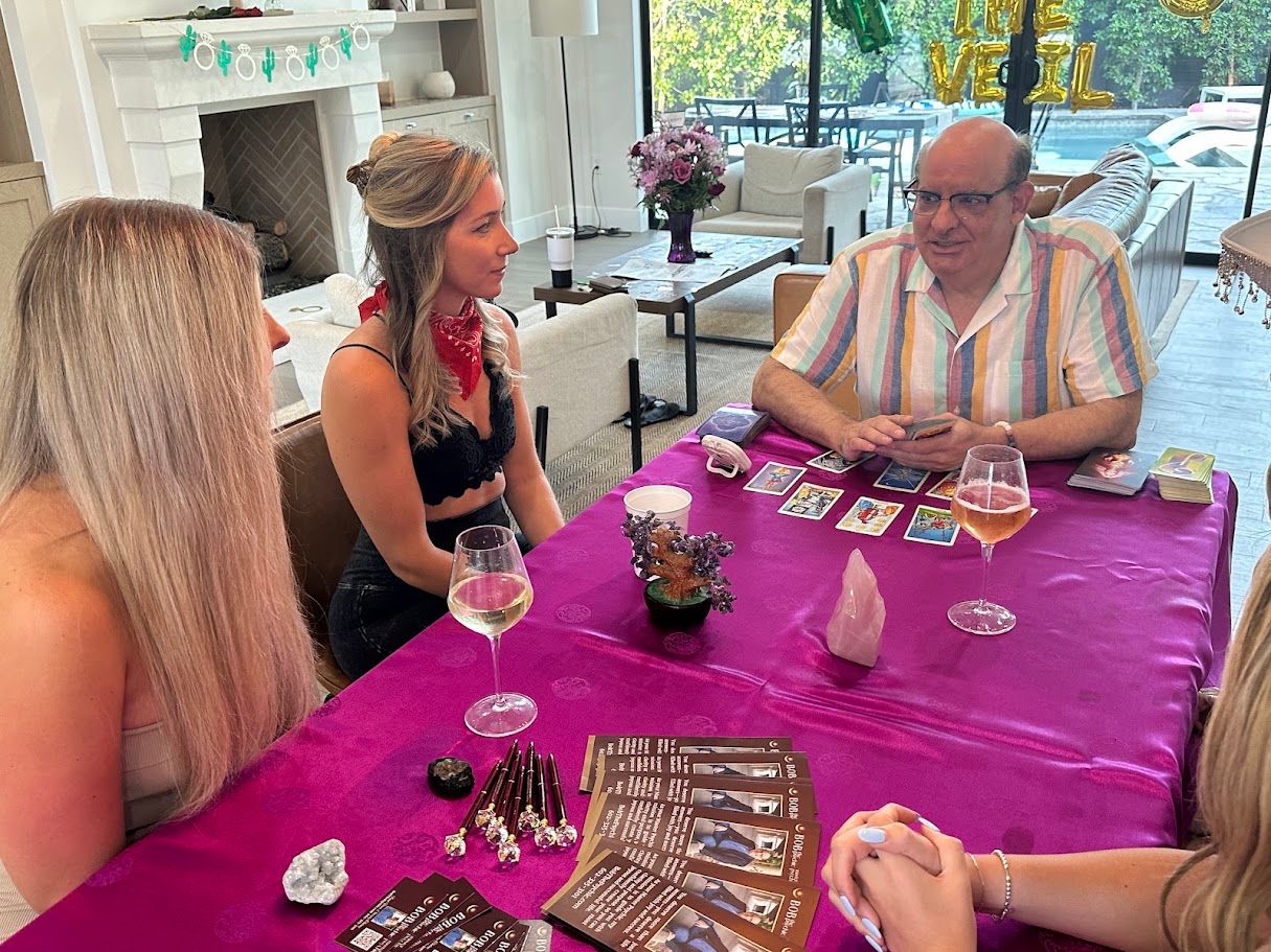 Tarot reading party in a bright modern living room by sliding glass doors and pool — a reader lays out tarot cards on a purple tablecloth with crystals, brochures and wine glasses while three women listen.