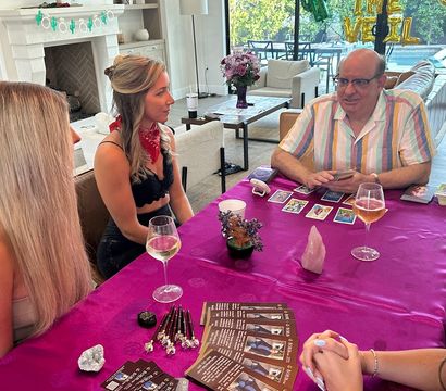 Tarot reading party in a bright modern living room by sliding glass doors and pool — a reader lays out tarot cards on a purple tablecloth with crystals, brochures and wine glasses while three women listen.
