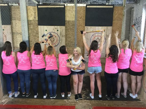 Group of women in matching pink tank tops at an indoor axe-throwing range, lined up with backs to camera and raising axes toward wooden bullseye targets while one woman smiles facing the camera.
