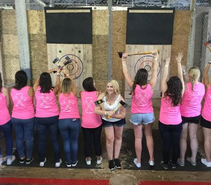 Group of women in matching pink tank tops at an indoor axe-throwing range, lined up with backs to camera and raising axes toward wooden bullseye targets while one woman smiles facing the camera.