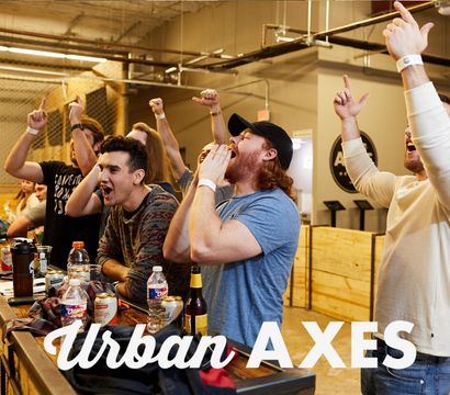 Group of friends cheering at an indoor axe-throwing venue, leaning on a wooden bar with drinks and raised hands in celebration.
