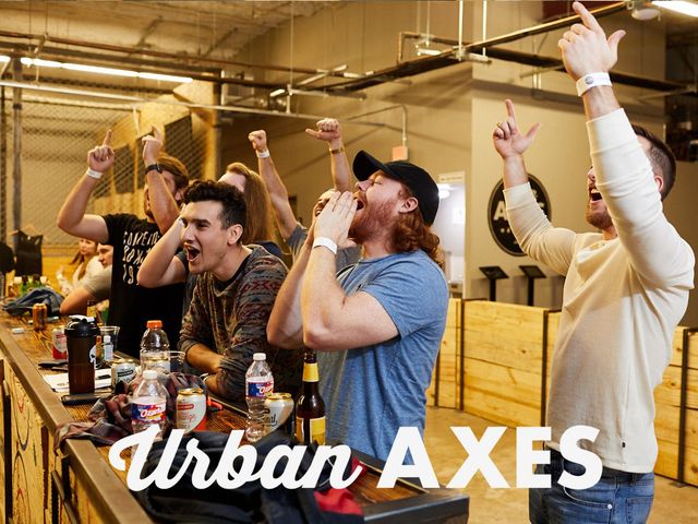 Group of friends cheering at an indoor axe-throwing venue, leaning on a wooden bar with drinks and raised hands in celebration.