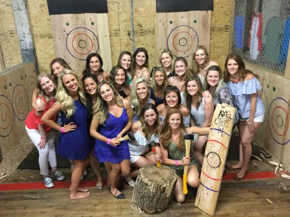 Group of smiling women posing at an indoor axe-throwing range with wooden targets, axes and a log stump — casual party photo.