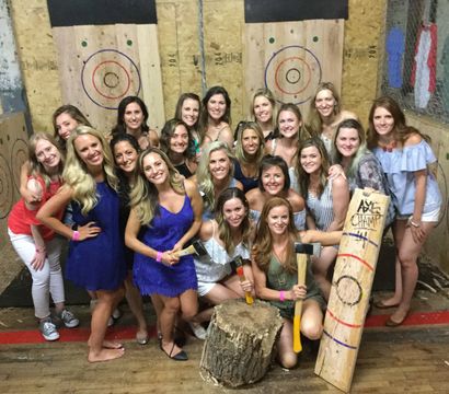 Group of smiling women posing at an indoor axe-throwing range with wooden targets, axes and a log stump — casual party photo.