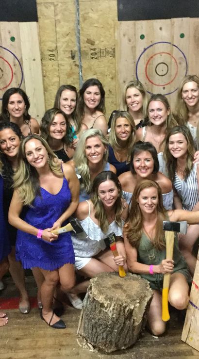 Group of smiling women posing at an indoor axe-throwing range with wooden targets, axes and a log stump — casual party photo.