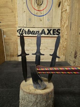 Three black throwing knives stuck tip-down in a round log stump before a painted bullseye on a wooden target, with a colorful striped-handled axe lodged in the stump in an indoor throwing lane.