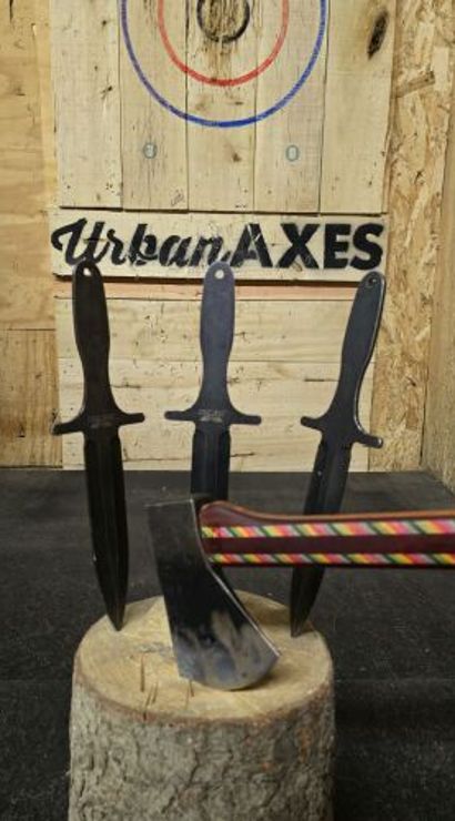 Three black throwing knives stuck tip-down in a round log stump before a painted bullseye on a wooden target, with a colorful striped-handled axe lodged in the stump in an indoor throwing lane.