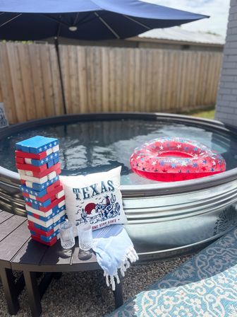 Backyard Texas stock-tank pool scene with a red, white and blue star-pattern inflatable ring floating, matching patriotic giant Jenga blocks stacked on a side table, a 'TEXAS' pillow, two clear glasses and a striped towel under a patio umbrella.