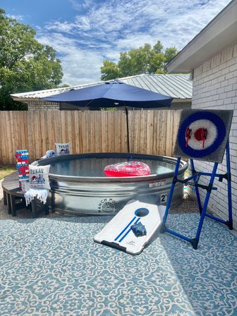 Sunny backyard patio with a round galvanized stock-tank pool under a navy umbrella, a red star-patterned inflatable ring floating, cornhole board on a blue patterned outdoor rug, an axe target board, and Texas-themed pillows by a wooden fence