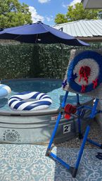 Sunny backyard stock-tank pool with a navy umbrella, striped blue-and-white inflatable float, and a freestanding Velcro dart target game on a patterned patio rug.