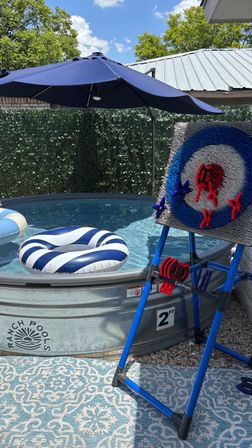 Sunny backyard stock-tank pool with a navy umbrella, striped blue-and-white inflatable float, and a freestanding Velcro dart target game on a patterned patio rug.