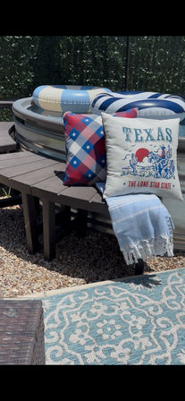 Texas patio with metal stock-tank pool, blue-and-white inflatable tubes, bench topped with a Texas-themed throw pillow and star-pattern cushion, striped towel draped over the seat, and a turquoise outdoor rug on gravel