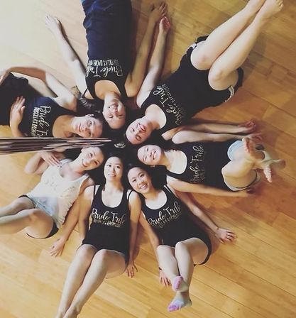 Eight women in matching 'Bride Tribe' tanks lying head-to-head in a star formation on a hardwood floor, smiling in a playful bachelorette party group photo